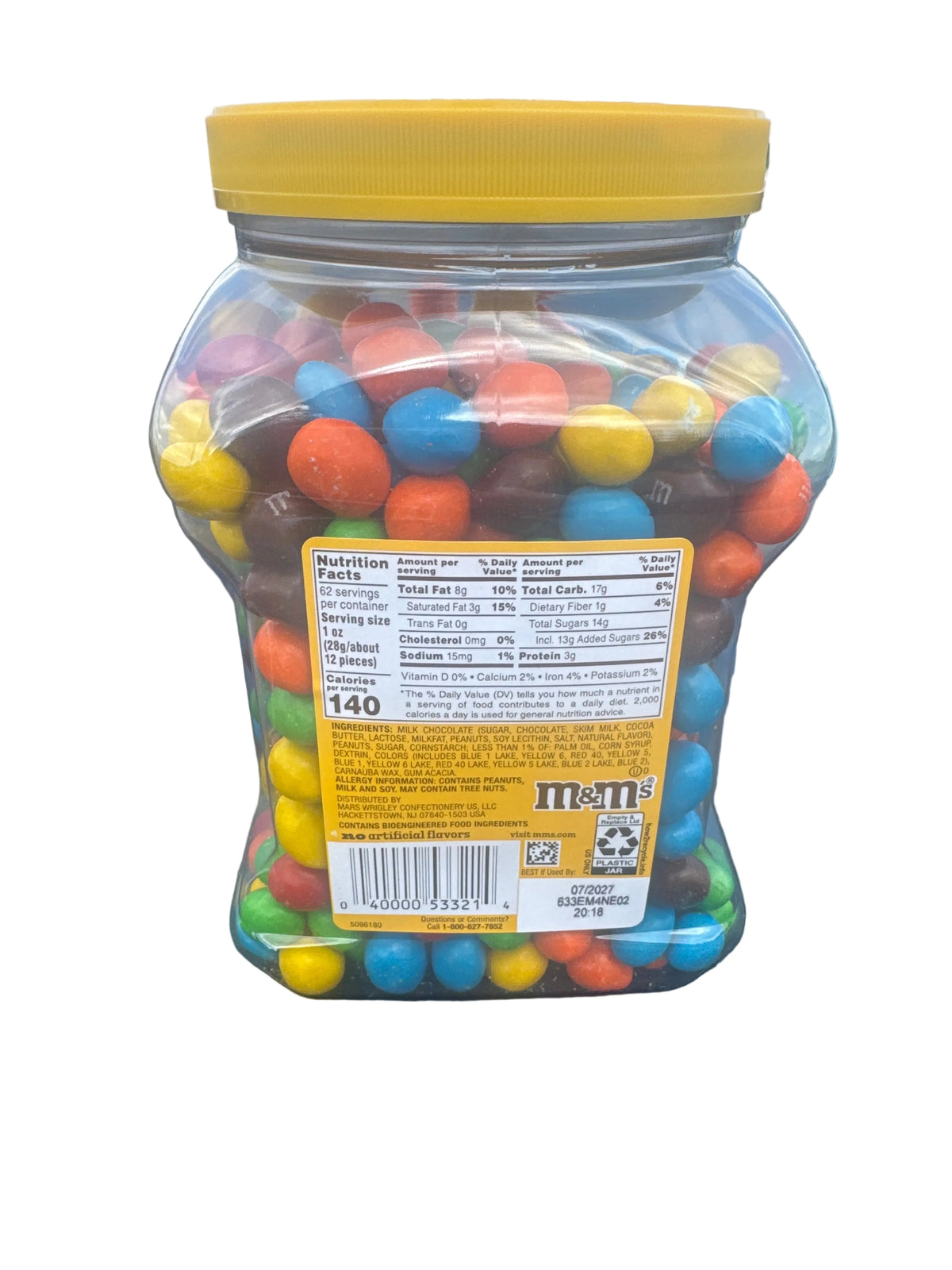 A large clear plastic jar filled with colorful M&M's candies, featuring a bright yellow lid and a yellow nutrition facts label on the back, photographed outdoors on the hood of a car.