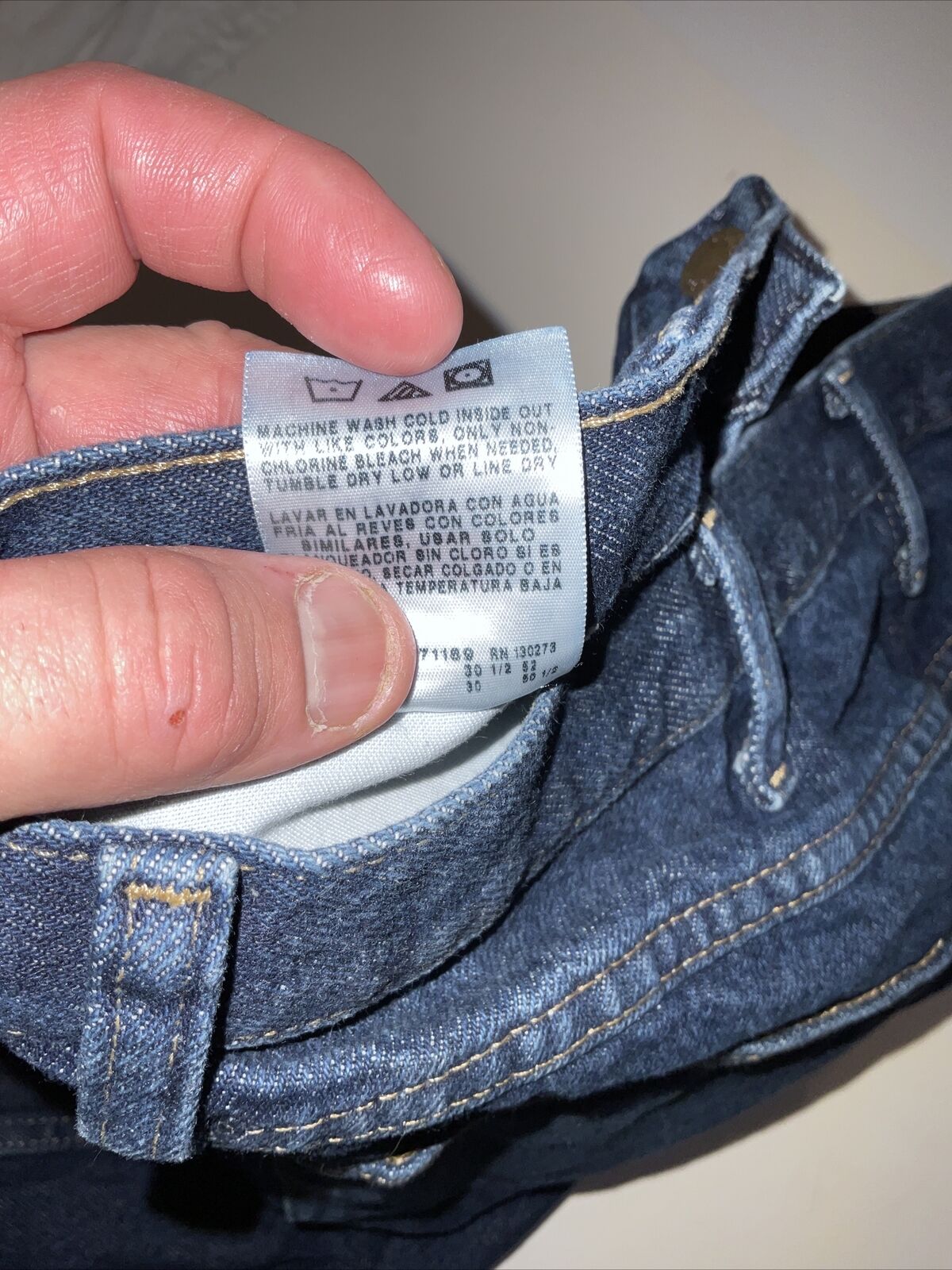 Hand holding care tag inside blue denim Wrangler relaxed fit jeans men waistband showing stitching and belt loop