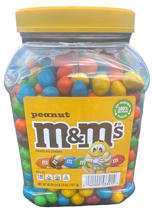A large clear plastic jar of M&M's peanut candy with a yellow lid and yellow front label, filled with colorful candy pieces, photographed outdoors on the hood of a dark car.