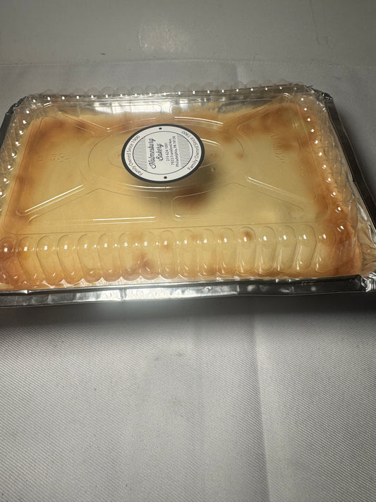 A square plastic clamshell container filled with a pale yellow custard or cream-colored dessert, photographed on a light gray fabric surface. A round white label is visible in the center.